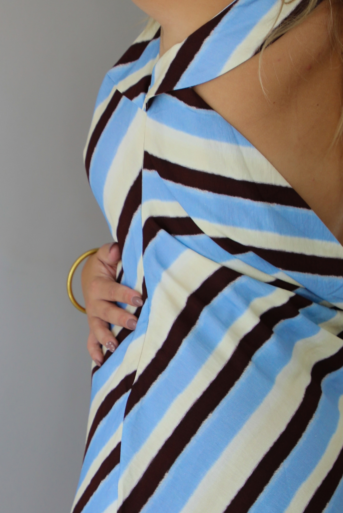 A woman wearing a cream, blue, and brown striped designed mini dress that has a high neckline with a mini length, standing indoors against a white wall. Th photo shows a closeup of the side of the dress, which has a zipper closure. 