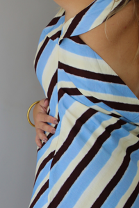 A woman wearing a cream, blue, and brown striped designed mini dress that has a high neckline with a mini length, standing indoors against a white wall. Th photo shows a closeup of the side of the dress, which has a zipper closure. 