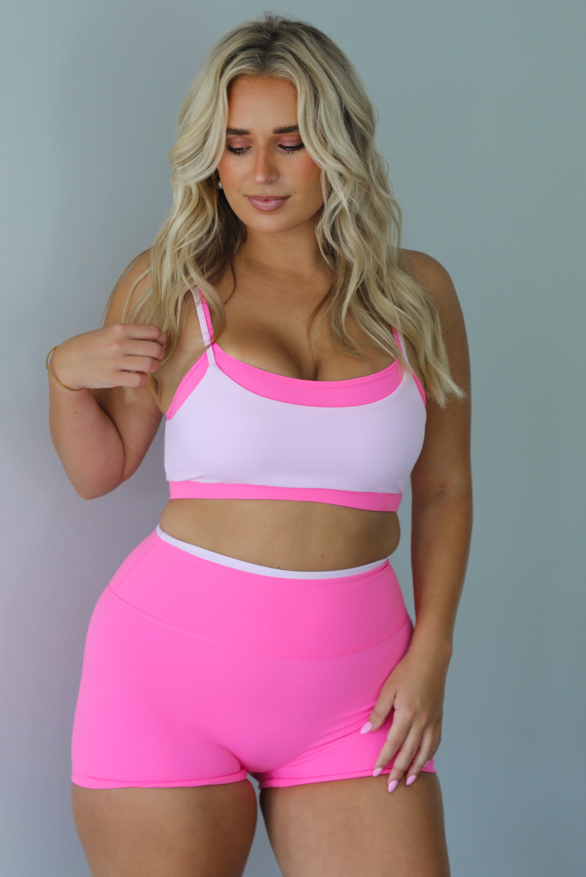 A woman modeling a bright pink and light pink two-piece activewear set consisting of a sports bra and matching high-waisted shorts, standing indoors against a white wall.