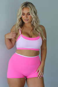A woman modeling a bright pink and light pink two-piece activewear set consisting of a sports bra and matching high-waisted shorts, standing indoors against a white wall.