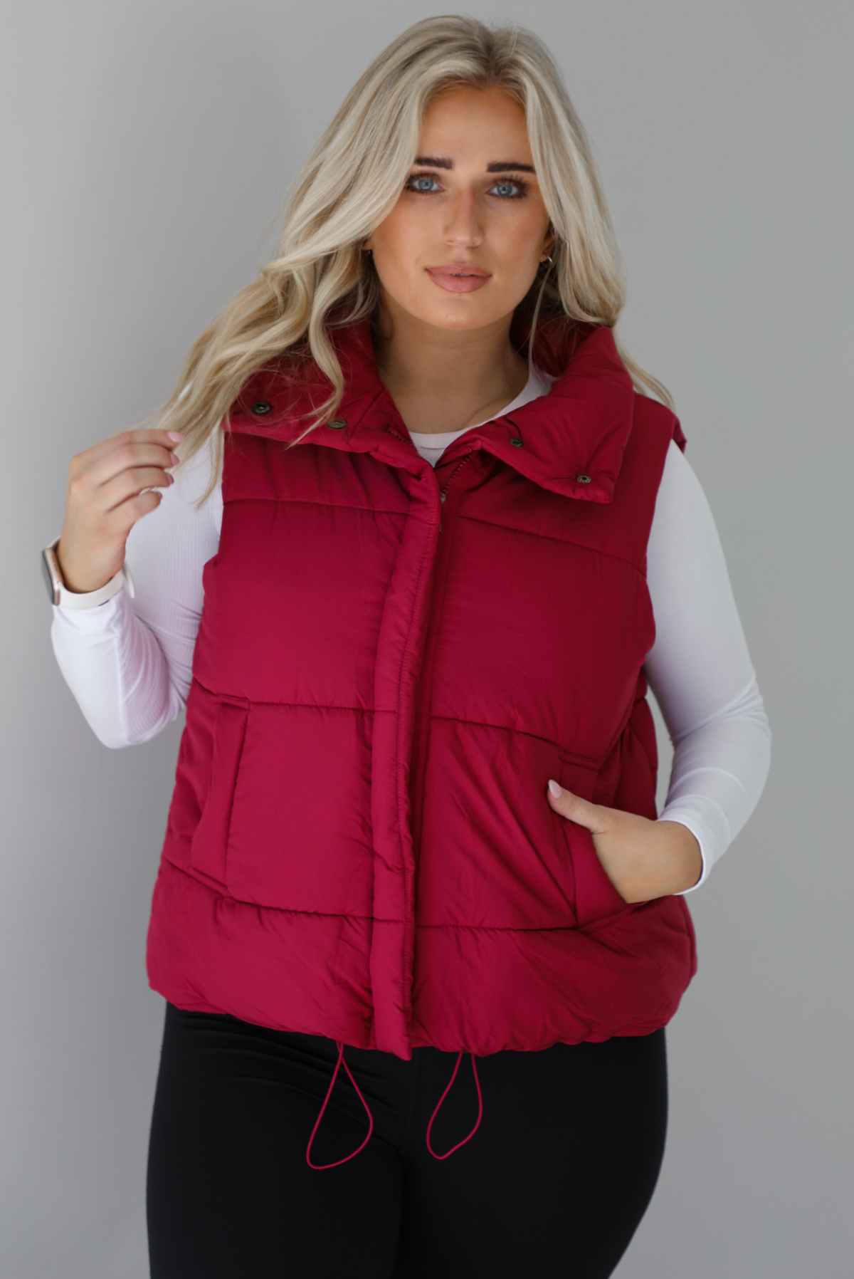 Woman wearing a wine red puffer vest, white long-sleeve shirt, and black leggings indoors against a white wall. The puffer vest has a collared design, functional button closures at the neckline, a zipper closure in the front, and functional pockets on either side.