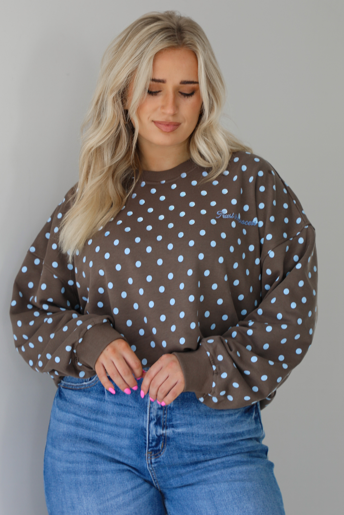 Woman wearing a brown sweater that has a blue polka dot print pattern throughout with blue embroidered writing on the left side that reads 'Trust The Process.' The sweater is paired with a medium wash denim. The photo shows a closeup of the sweater, which is folded under at the hem to give a cropped look. The photo is taken indoors against a white wall.