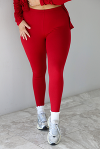 Woman wearing a red two piece set that features an oversized long sleeved top and fitted leggings. The photo shows a closeup of the pants that are paired with high white ankle socks and silver sneakers. The photo is taken indoors against a white wall with white marble flooring.