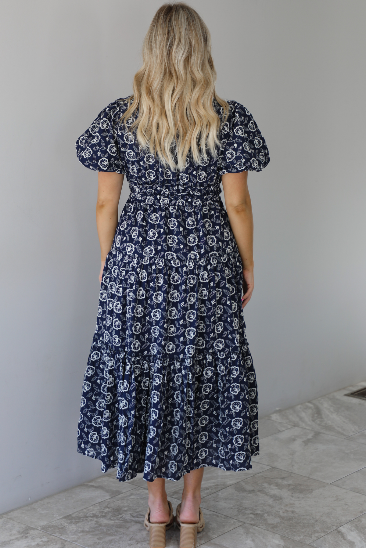 Woman wearing a navy dress that has a white embroidered floral print pattern throughout, a cinched waistline, and flowy maxi length skirt. The photo shows the back of the dress and is taken in front of a white wall with white marble flooring. 
