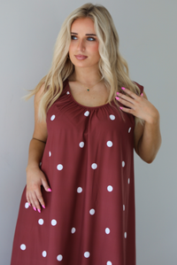 Woman wearing a red brown maxi length dress that has a white polka dot print pattern throughout. The photo shows a closeup of the upper half of the dress. The photo is taken indoors against a white wall.