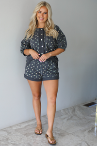 Woman wearing a charcoal with a multi-colored floral two piece set featuring an oversized top with puffed sleeves and shorts indoors against a white wall with white marble flooring. The top is tucked into the front of the shorts.