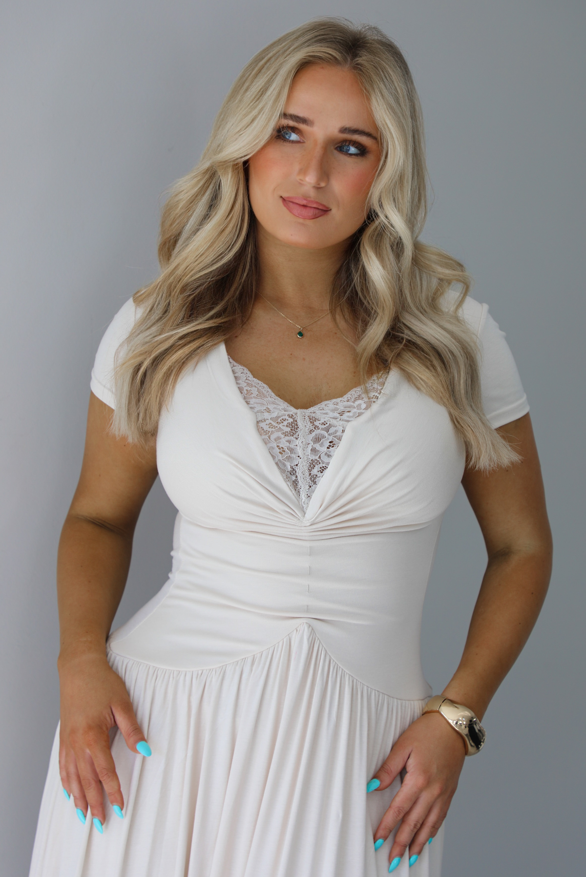 Woman wearing a light sand colored maxi length dress that has a fitted bodice with lace detailing in the center of the bust, a drop waist design, short sleeves, and a flowy skirt. The photo is taken indoors against a white wall and shows a closeup of the bodice of the dress. 