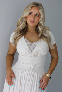 Woman wearing a light sand colored maxi length dress that has a fitted bodice with lace detailing in the center of the bust, a drop waist design, short sleeves, and a flowy skirt. The photo is taken indoors against a white wall and shows a closeup of the bodice of the dress. 