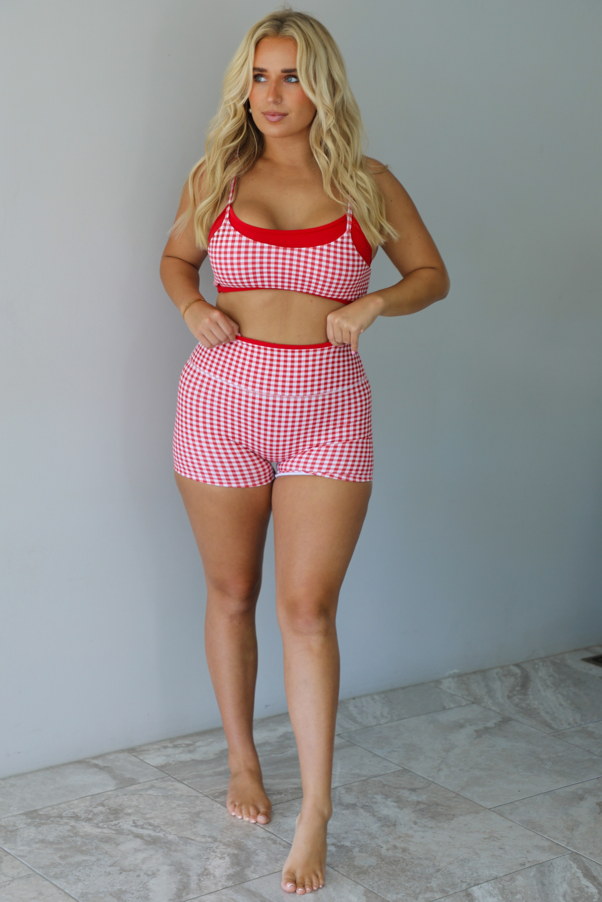 A woman modeling a red and white gingham two-piece athletic set featuring a sports bra and high waisted biker shorts with solid red trim indoors against a white wall with white marble flooring. 