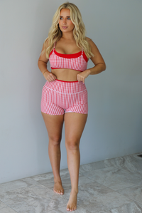 A woman modeling a red and white gingham two-piece athletic set featuring a sports bra and high waisted biker shorts with solid red trim indoors against a white wall with white marble flooring. 