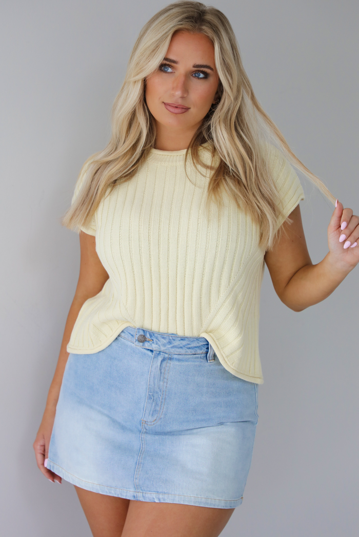 Woman wearing a pale yellow short sleeved kit top with a light wash denim skort. The photo is taken indoors against a white wall.