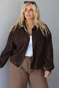 A woman wearing a dark brown suede jacket with a white tank and mocha colored mid-to-low rise pants, standing indoors against a white wall.