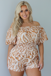 Woman wearing a tan and white romper that has off the shoulder short puffed sleeves, a cinched waistline, and a repeating leaf pattern indoors against a white wall.