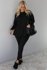 Woman wearing a black two piece set that features an oversized long sleeved top and fitted leggings. The set is paired with a black vegan leather shoulder bag and black slide on slippers. The photo is taken indoors against a white wall with white marble flooring.