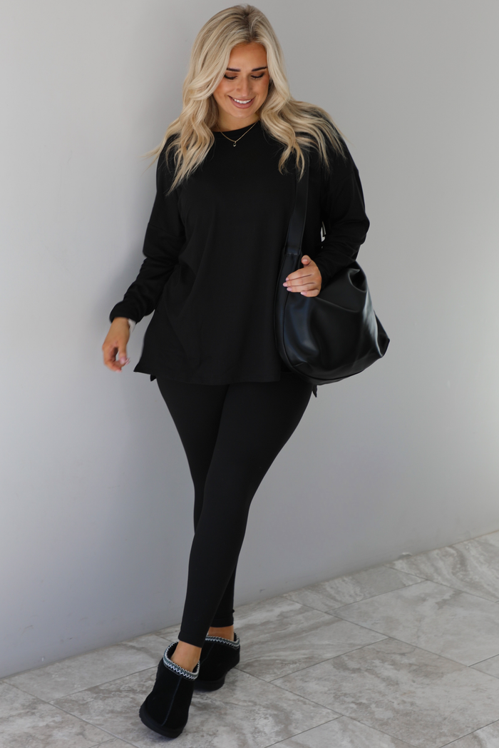 Woman wearing a black two piece set that features an oversized long sleeved top and fitted leggings. The set is paired with a black vegan leather shoulder bag and black slide on slippers. The photo is taken indoors against a white wall with white marble flooring.