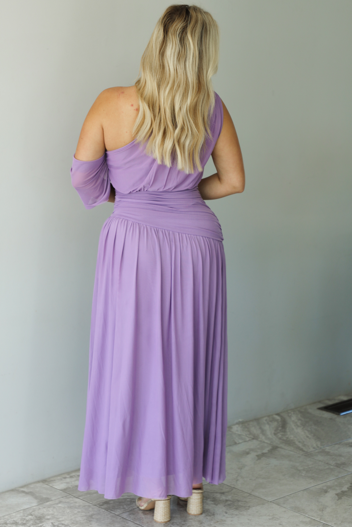 Woman wearing a lavender formal gown featuring a one strap that has extra mesh material that goes to the other arm and drapes down, has a fitted drop waist, and a long, maxi length skirt. The photo is taken indoors against a white wall with white marble flooring. The photo shows the back of the dress. 