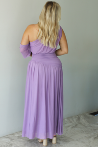 Woman wearing a lavender formal gown featuring a one strap that has extra mesh material that goes to the other arm and drapes down, has a fitted drop waist, and a long, maxi length skirt. The photo is taken indoors against a white wall with white marble flooring. The photo shows the back of the dress. 
