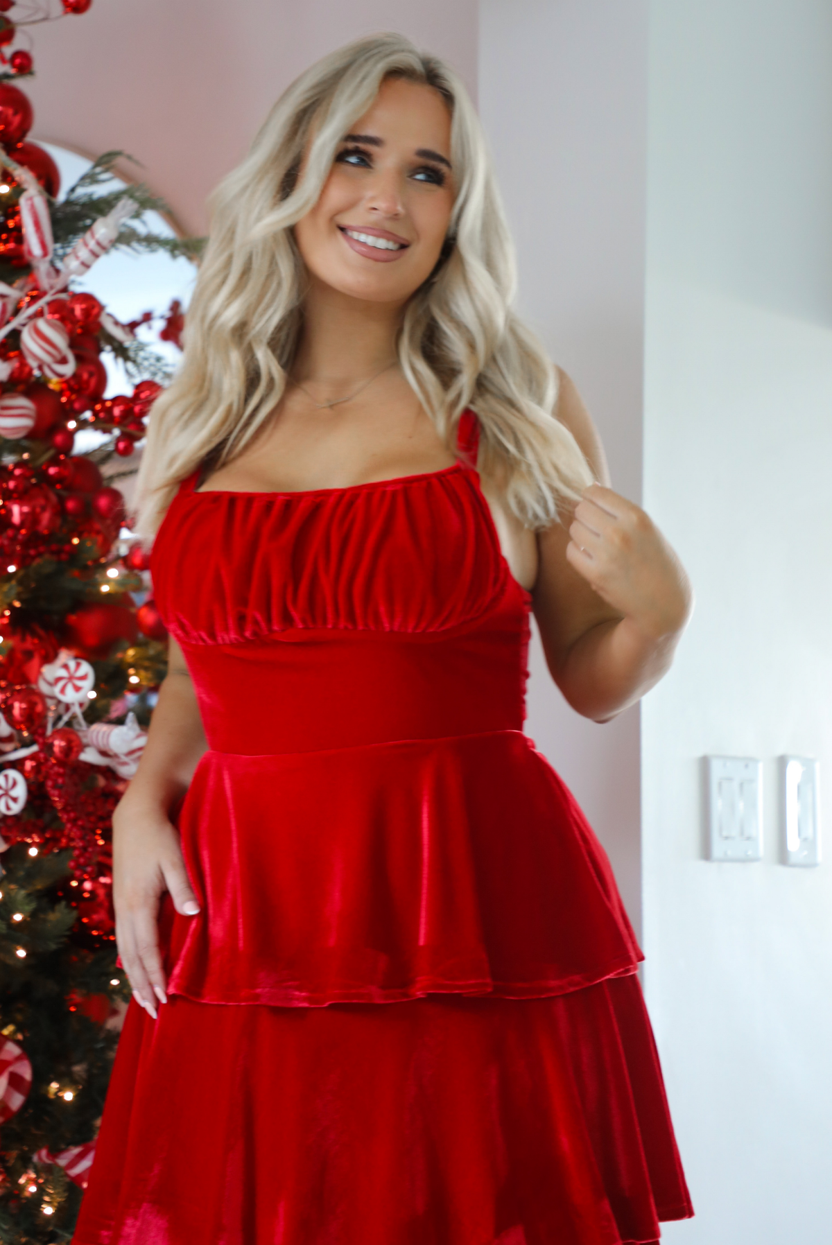 Close up of a woman model wearing a red velvet textured ruffled maxi dress. The dress has adjustable straps, a fitted waistline, and a ruffled skirt.  The photo showcases the bodice and the supper half of the dress. The photo is taken inside a modern home with a decorated Christmas tree in the back. 