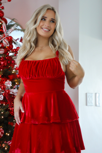 Close up of a woman model wearing a red velvet textured ruffled maxi dress. The dress has adjustable straps, a fitted waistline, and a ruffled skirt.  The photo showcases the bodice and the supper half of the dress. The photo is taken inside a modern home with a decorated Christmas tree in the back. 