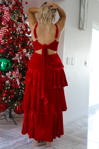 Back of a woman model wearing a red velvet textured ruffled maxi dress. The dress has adjustable straps, a fitted waistline, and a ruffled skirt. The photo showcases the adjustable straps in the back, a tie closure in the back, and an elastic waistline. The photo is taken inside a modern home with a decorated Christmas tree in the back. 