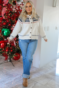 Woman model wearing a white, navy, and red patterned cardigan and jeans standing in front of a decorated Christmas tree.The cardigan has an oversized fit, functional button closures in the front, and is super soft and comfortable. 