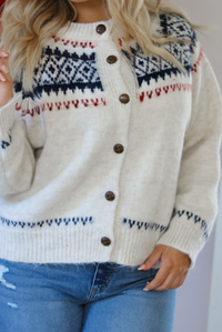Close up of a woman model wearing a white, navy, and red patterned cardigan and jeans standing in front of a decorated Christmas tree.The cardigan has an oversized fit, functional button closures in the front, and is super soft and comfortable. 