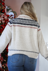 Back of a woman model wearing a white, navy, and red patterned cardigan and jeans standing in front of a decorated Christmas tree.The cardigan has an oversized fit, functional button closures in the front, and is super soft and comfortable. 