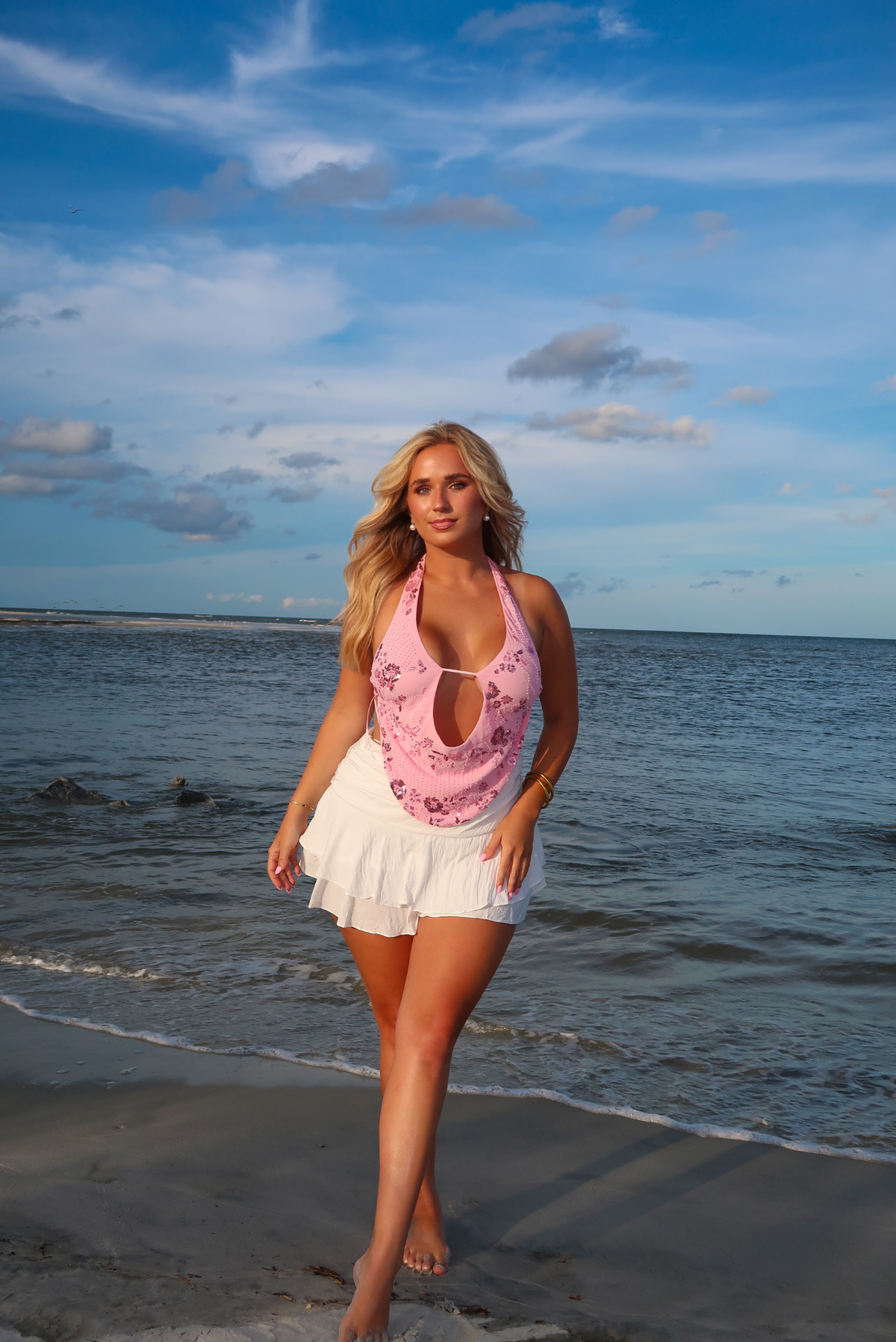 A woman stands on a beach shoreline wearing a pink floral halter top with a white ruffled skirt, against a blue ocean and cloudy sky background.