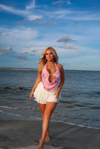 A woman stands on a beach shoreline wearing a pink floral halter top with a white ruffled skirt, against a blue ocean and cloudy sky background.