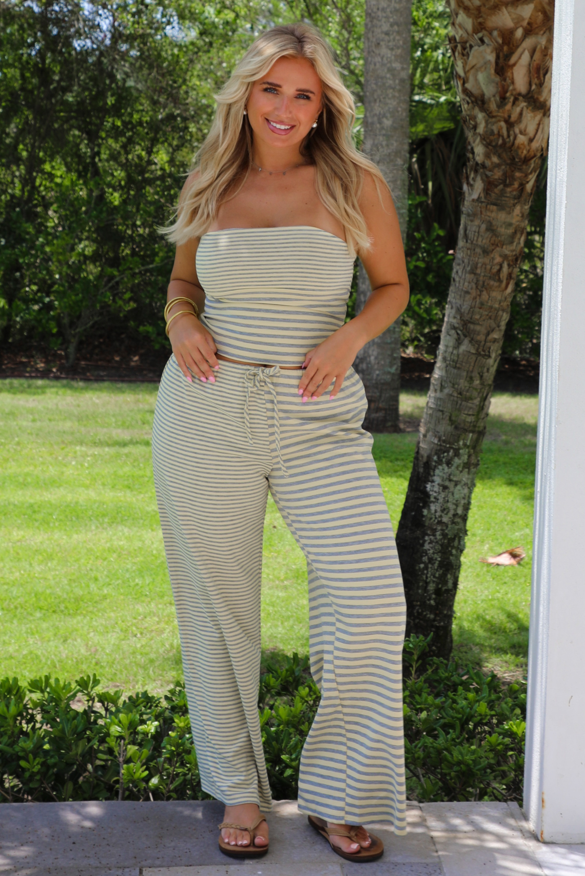 A woman wearing a pale yellow and heather grey striped two piece set with a sleeveless tube top and wide-leg pants, standing outdoors on a patio with a green lawn and trees in the background.