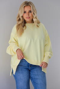Woman wearing a pale yellow oversized sweater that's untucked and paired with dark wash denim. The photo is taken indoors against a white wall.