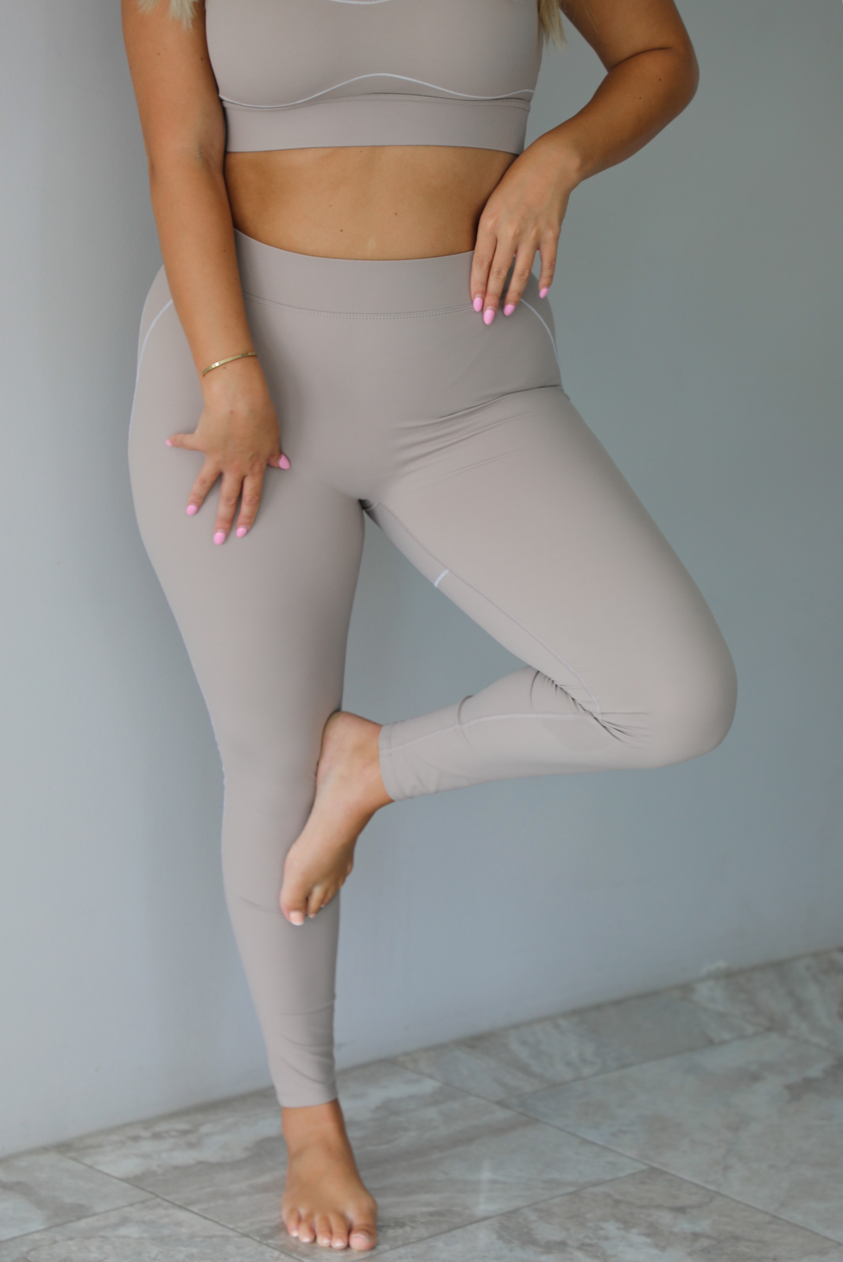 A woman modeling a matching tan activewear set consisting of a sports bra and high-waisted leggings with white stitching details, standing indoors against a white wall.