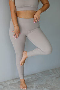 A woman modeling a matching tan activewear set consisting of a sports bra and high-waisted leggings with white stitching details, standing indoors against a white wall.