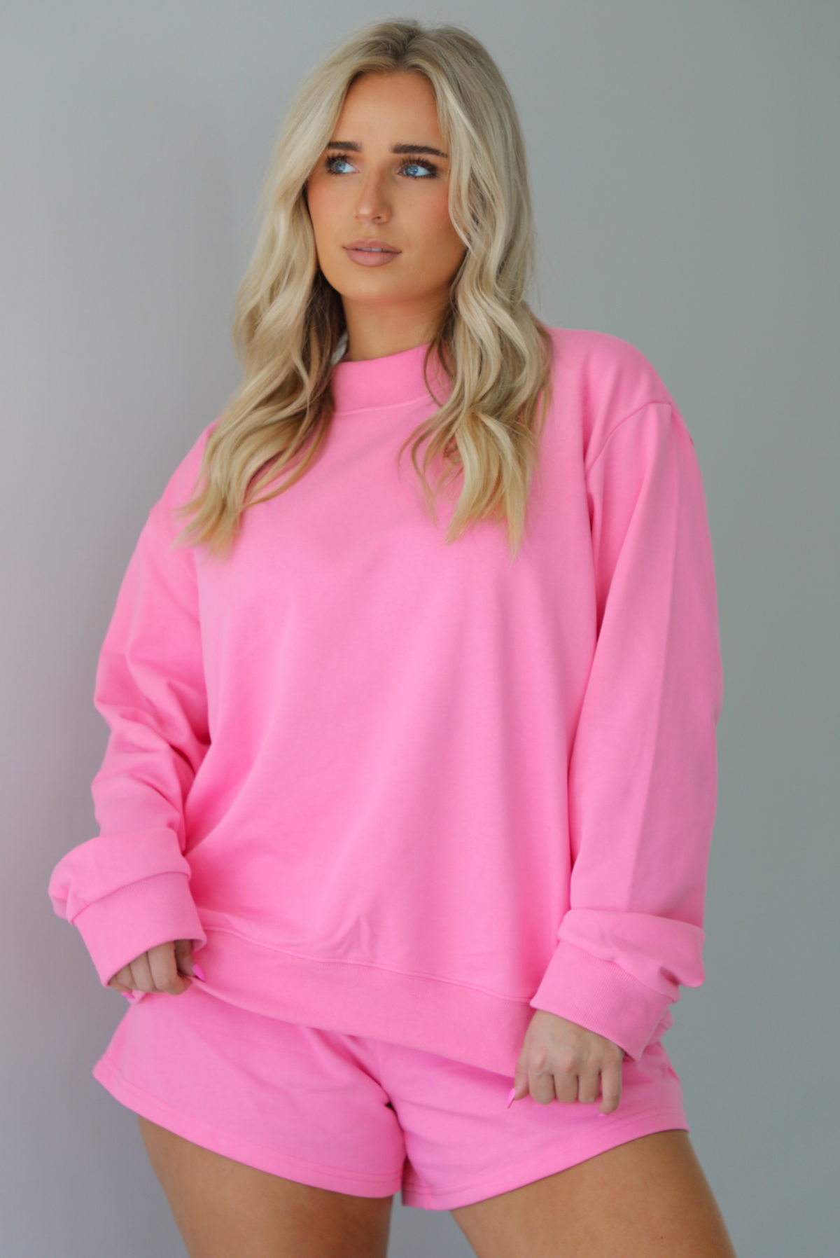 Woman wearing a hot pink two piece set featuring an oversized sweatshirt and high waisted shorts. The photo is taken indoors against a white wall and shows a closeup of the top of the set, which is untucked to show the full length. 