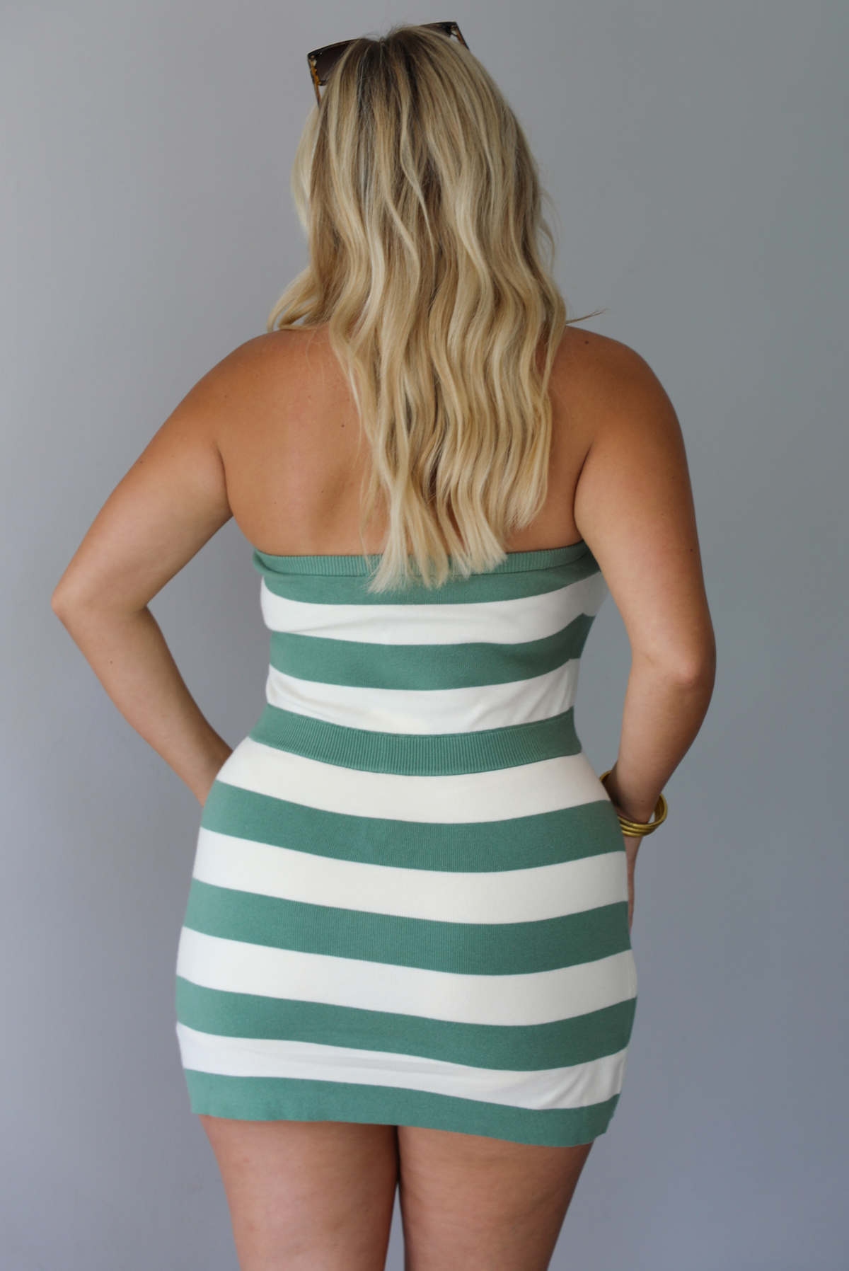 A woman shown from behind modeling a strapless mini dress with horizontal sage green and white stripes, standing indoors against a white wall.
