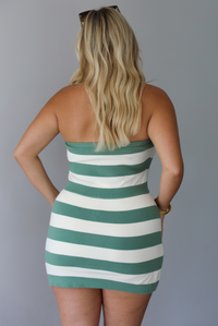 A woman shown from behind modeling a strapless mini dress with horizontal sage green and white stripes, standing indoors against a white wall.