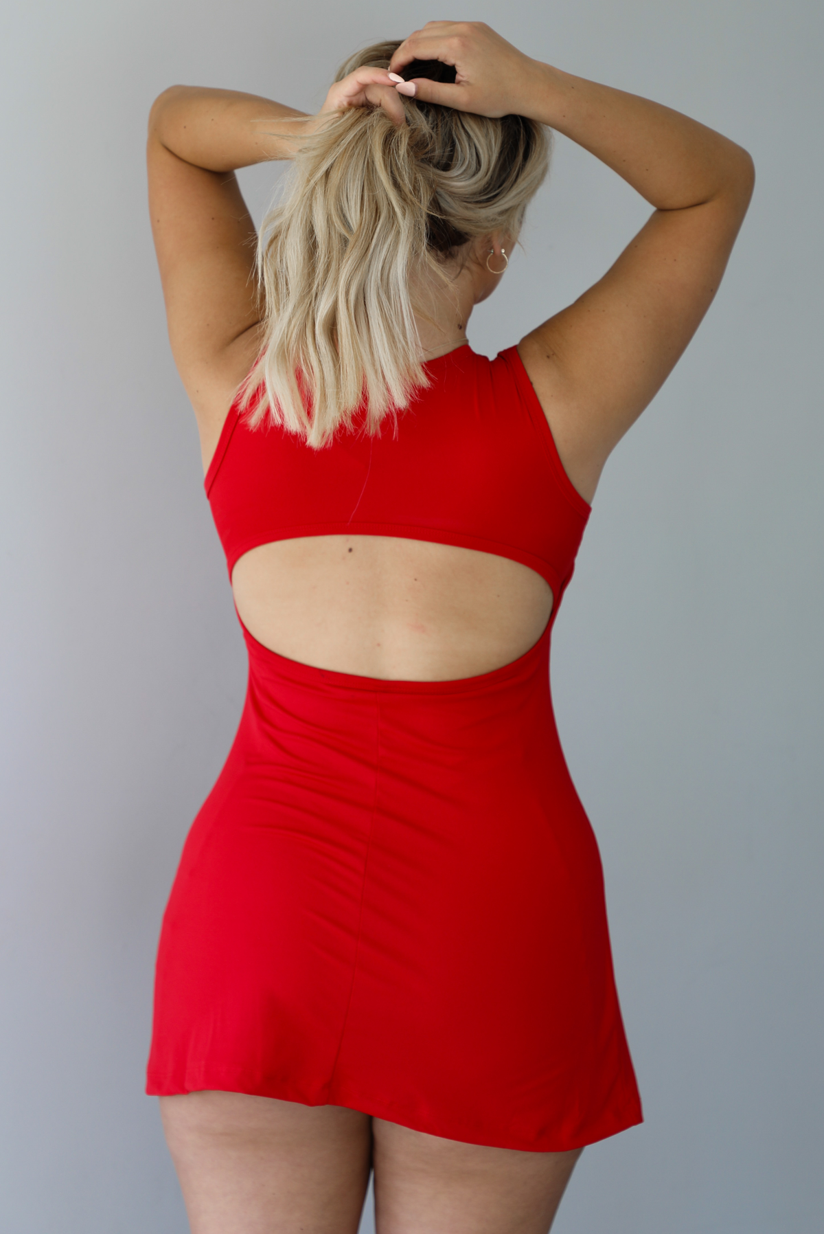 Woman wearing a red athleisure dress with a rounded neckline, thicker material, and a mini length indoors against a white wall. The photo shows the back of the dress, which has a cut out detail. 