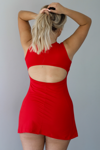 Woman wearing a red athleisure dress with a rounded neckline, thicker material, and a mini length indoors against a white wall. The photo shows the back of the dress, which has a cut out detail. 