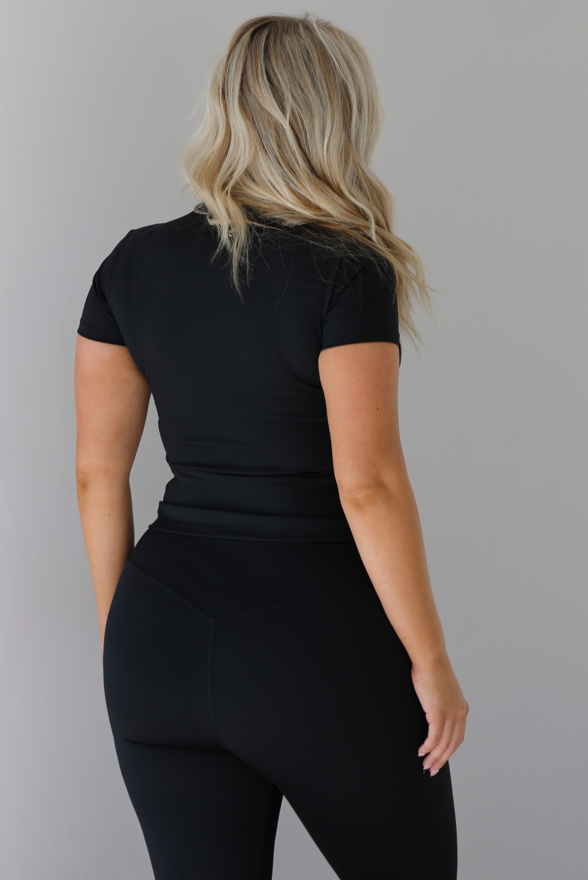 Woman wearing a fitted two piece black set featuring a short sleeved top and fitted leggings. The photo shows a closeup of the back of the top of the set and the top of the back of the pants. The photo is taken indoors against a white wall.