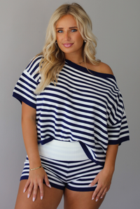 A woman wearing a navy and white horizontally striped off-the-shoulder top and matching striped shorts, standing indoors against a white wall.