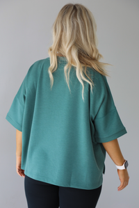 Woman wearing a teal oversized t-shirt with black leggings. The photo shows the back of the tee. The photo is taken in front of a white wall indoors. 