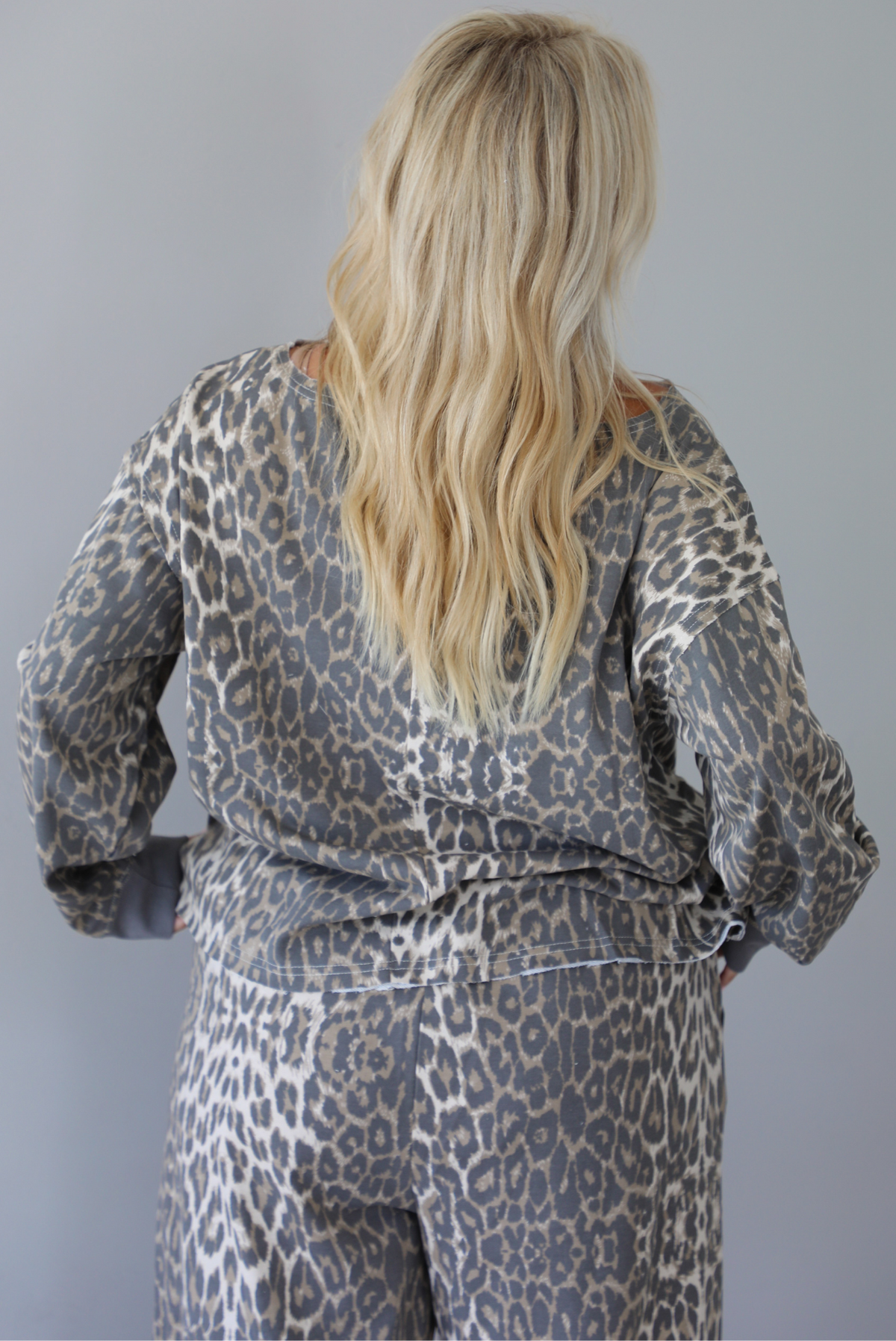 A woman wearing a leopard patterned two piece set featuring an oversized top with a front pocket detail and mid-rise oversized pants, standing indoors against a white wall. The photo shows a closeup of the back of the top. 
