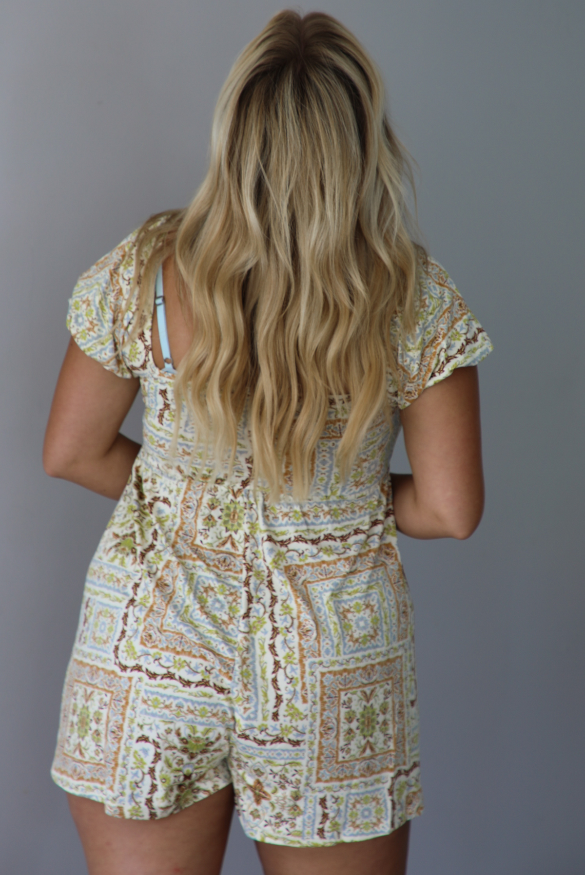 A woman wearing a cream-colored short-sleeve romper with a beige and brown geometric pattern, standing indoors against a white wall. Th photo shows the back of the romper. 