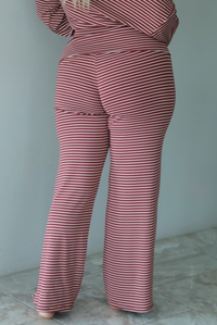 A woman shown from behind wearing a maroon and white striped two piece matching set featuring a slouchy off the shoulder long sleeved top and mid-rise, folded over waistline wide leg pants, standing indoors against a white wall with white marble flooring. The photo shows a close up of the back of the pants. 