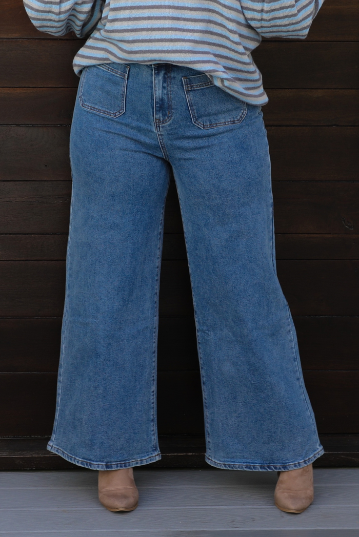 A woman wearing medium wash flared leg denim with small pockets on the front, in front of a dark wooden door. 