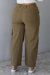 A closeup of a woman shown from behind wearing olive khaki pants with a mid-rise waistline, pocket details, with a wide leg design, standing indoors against a white wall with white marble flooring. The back of the pants has functional back pockets. 