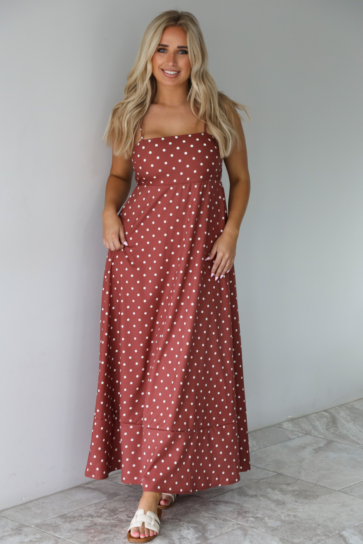 Woman wearing a spaghetti strap, maxi length dress that has a rust color throughout with a white polka dot pattern. The photo is taken indoors against a white wall with white marble flooring.