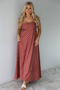 Woman wearing a spaghetti strap, maxi length dress that has a rust color throughout with a white polka dot pattern. The photo is taken indoors against a white wall with white marble flooring.