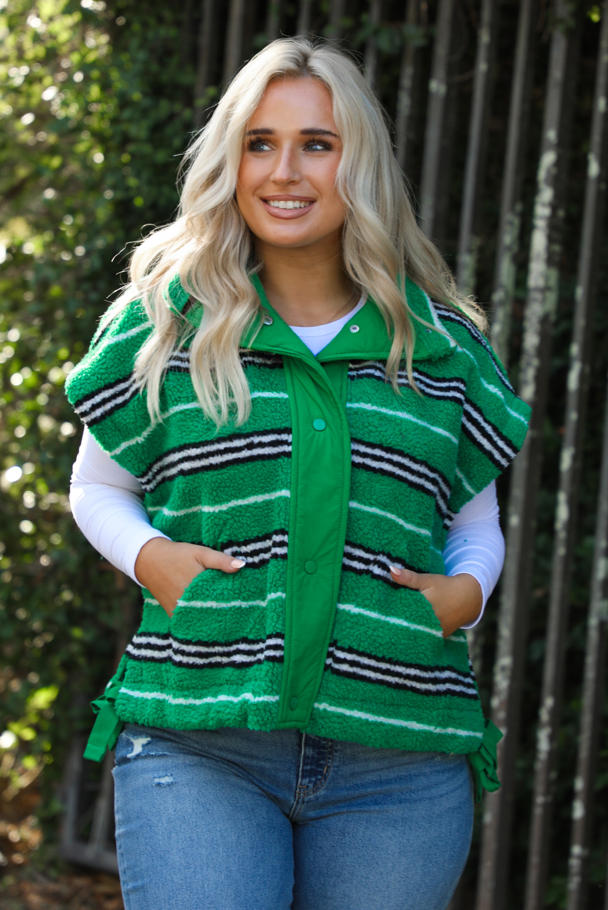 Woman model wearing a green, black, and white striped vest that has a sherpa texture to it, functional button closures in the front, and tie closures on either side, It is paired with a white long sleeved top, a medium wash pair of denim jeans, and tan ugg boots. The photo is taken outdoors with a metal gate and greenery in the back. The vest is shown close up, displaying the buttons closed and the pockets in use on either side.
