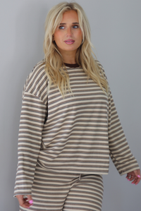 A woman wearing a sand colored matching two piece set with a brown striped pattern throughout featuring an oversized long sleeved top and oversized slouchy wide leg pants, standing indoors against a white wall. The photo shows a closeup of the top untucked to show the full length. 